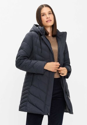 Woman wearing a dark gray padded hooded winter coat over a brown sweater and black pants, adjusting the coat’s zipper.