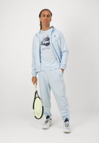 Gai zils sporta tērps, kas izgatavots no mīkstas auduma, ar grafisko Lacoste logo uz krekla. Subjekts tur tenisa raketi un valkā sporta apavus.