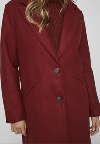 Manteau en laine bordeaux avec col cranté, deux boutons et poches latérales. Présente une texture lisse et une coupe cintrée.