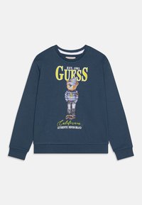 Granatowy sweter z niedźwiedziem w czapce marynarskiej i w paski koszulce, z napisem "GUESS EST. 1981 California Authentic Denim Brand" z przodu.