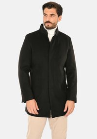 Cappotto nero in lana con colletto alto, taglio dritto e tasche laterali. Presenta una texture liscia e un design semplice e moderno.