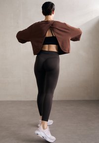 Brauner, kurz geschnittener Sweatshirt mit offenem Rücken, schwarzem Sport-BH, dunklen Leggings und hellpinkfarbene Turnschuhe. Glatte Textur, figurbetontes Design.