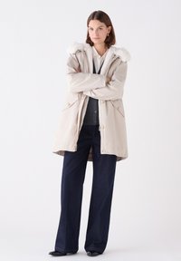 Beige parka met een bontgevoerde capuchon, voorzien van knopen en zakken, gecombineerd met donkerblauwe wijde jeans en zwarte schoenen.