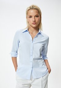 Lichtblauwe blouse met lange mouwen, knoopsluiting aan de voorkant, kraag en opgerolde manchetten; gladde stof met een iets getailleerde pasvorm en een gebogen zoom.