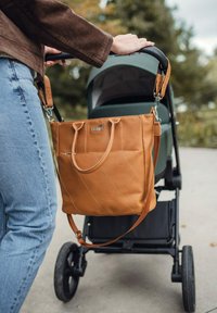 Kožená taška na rameno v barvě opálené kůže s diagonálním prošíváním a přední kapsou, zavěšená na rukojeti kočárku vedle osoby v džínách.