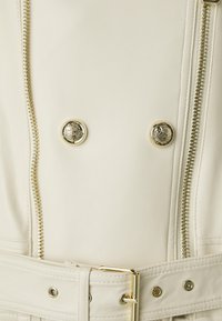 Veste en cuir synthétique crème avec zip et boutons dorés, dotée d'une taille ceinturée et d'un design croisé. Texture lisse, forme structurée.