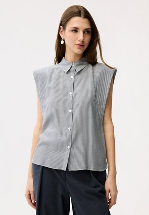 Ung kvinde iført ærmeløs gråstribet skjorte med knapper og mørke bukser, der kigger mod venstre mod en ensfarvet baggrund.