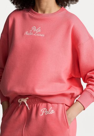 Persona que lleva un conjunto a juego de sudadera y pantalones deportivos de color coral rosa con los logotipos blancos bordados de "Polo Ralph Lauren" y "Polo". Manos en los bolsillos.