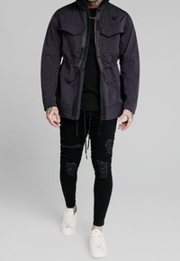 Lilla letvægtsjakke med to brystlommer og snoretræk i taljen, parret med slidte sorte skinny jeans og hvide sneakers.