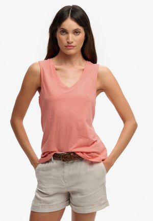 Vrouw met lang bruin haar, gekleed in een koraalkleurige mouwloze top met V-hals en beige korte broek met een gevlochten bruine riem, staande met handen in de zakken.