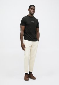 Homem preto em pé, vestindo uma t-shirt preta da Armani Exchange, calças em tom creme, meias pretas e ténis castanhos, contra um fundo claro e simples.