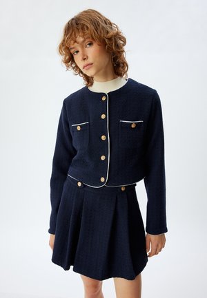 Giacca navy a trama con bottoni dorati e piping bianco, abbinata a una gonna plissettata coordinata. Entrambi i capi presentano tasche frontali.