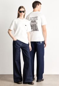 Άσπρα βαμβακερά t-shirts με γραφικό της Lacoste στην πλάτη, συνδυασμένα με φαρδιά μπλε τζιν παντελόνια. Το μοντέλο φοράει σκούρα γυαλιά ηλίου και καφέ παπούτσια.