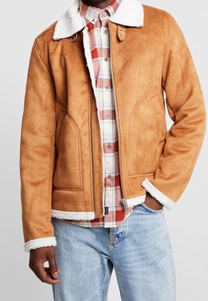 Homme portant une veste en peau lainée beige avec col et poignets en fourrure blanche, chemise à carreaux rouges et blancs, et jean bleu clair, main dans la poche.