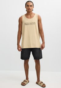 Hombre con el cabello trenzado, vestido con una camiseta sin mangas beige con el texto "White Peak", pantalones cortos negros y sandalias beige, posando frente a un fondo blanco.