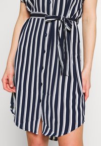 Abito a righe blu navy e bianche con bottoni, cintura in vita, maniche corte e orlo curvo. Presenta una texture di tessuto leggero e liscio.