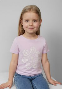 Lichtroze t-shirt met korte mouwen, versierd met een witte bloemenpatroon. Het kind draagt blauwe denim jeans en zit op een vlakke ondergrond.