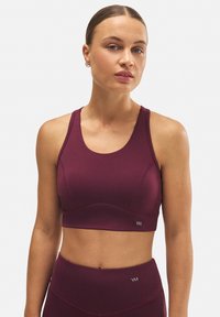 Sostenedor deportivo burdeos con escote redondeado, tirantes anchos y textura de punto. Presenta un logo en el panel inferior izquierdo.