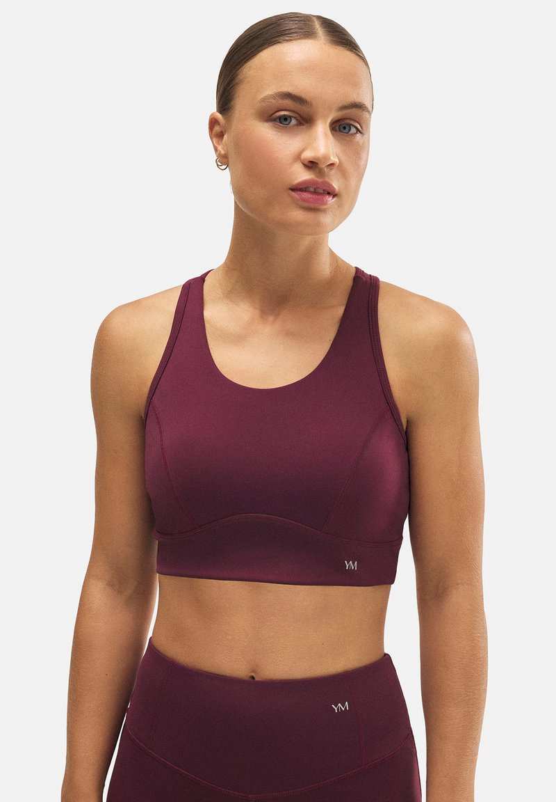 Sostenedor deportivo burdeos con escote redondeado, tirantes anchos y textura de punto. Presenta un logo en el panel inferior izquierdo.