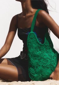 Vihreä virkattu kassi kukkakuviolla, jossa on himmeä pinta ja kaksi tukevasti käsikahvaa, vaaleaa taustaa vasten.