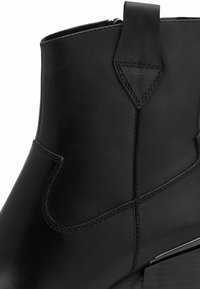 Bottine en cuir noir avec un talon plat, présentant une texture lisse, des coutures décoratives et une fermeture éclair sur le côté.