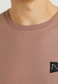 Krátký rukáv, mauve tričko s kulatým výstřihem, se černou trojúhelníkovou nášivkou na levém prsu a žebrovanou texturou.