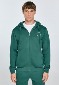 Tmavě zelená mikina na zip vyrobená z měkké tkaniny, s kapucí na šňůrku, dvěma předními kapsami a malým bílým logem na hrudi.