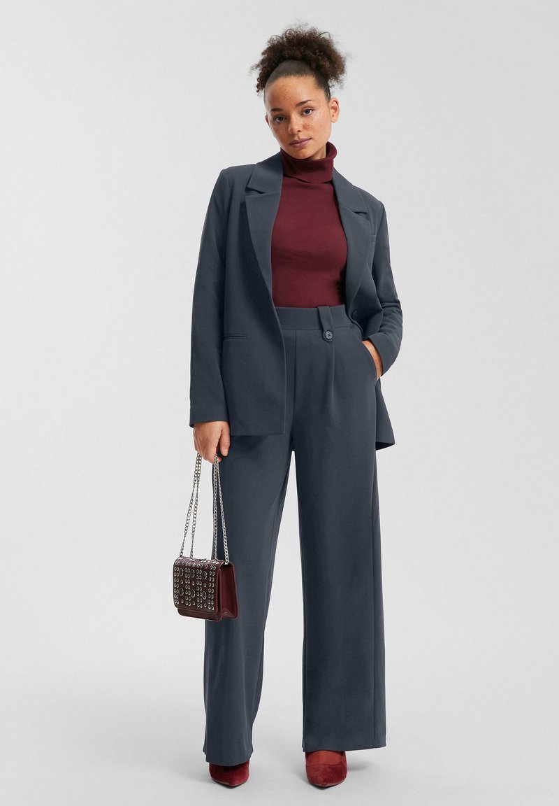 Completo grigio con blazer su misura e pantaloni a vita alta. Collo alto color bordeaux sotto, borsa bordeaux e tacchi abbinati.