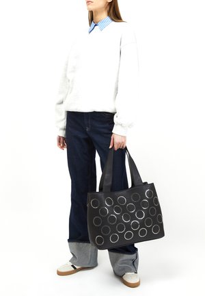 Zwarte draagtas met cirkelvormige metalen studs, gladde textuur, dubbele handvatten en een gestructureerde vorm. Draagt bij casual blauwe en grijze kleding.