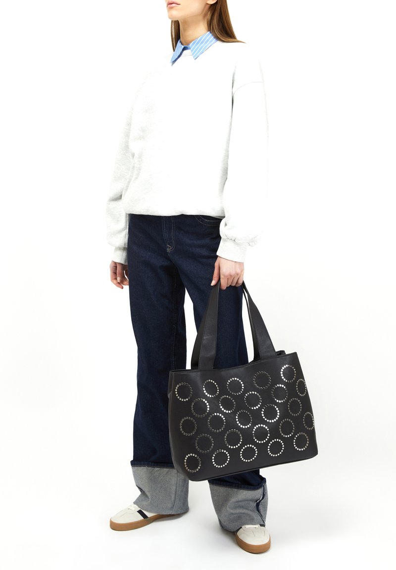 Borsa tote nera con rivetti metallici circolari, texture liscia, doppi manici e forma strutturata. Indossata con abbigliamento casual blu e grigio.