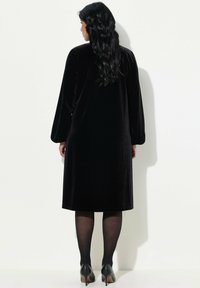 Robe en velours noir à manches longues, longueur genou et coupe décontractée. Associée à des chaussures à talons hauts noires et des collants transparents noirs.