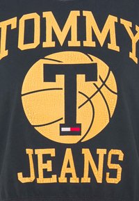 Černá látka má velký prasklý žlutý potisk s nápisem "TOMMY" nahoře a "JEANS" dole, který obsahuje grafiku basketbalového míče a malý červený, bílý a modrý akcent.