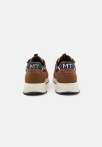 Brune semskede treningssko med en polstret hvit såle og en mørk blå hælflik med "MT"-merke i hvitt. Bakfra, minimalistisk design.