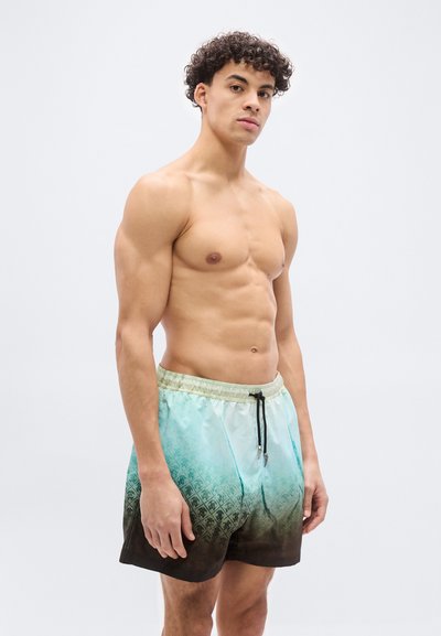 Γυμνόστηθος νεαρός με σγουρά μαλλιά που φοράει μαγιό σε ανοιχτό μπλε και μαύρο διαβάθμιση, στέκεται μπροστά σε λευκό, απλό φόντο.