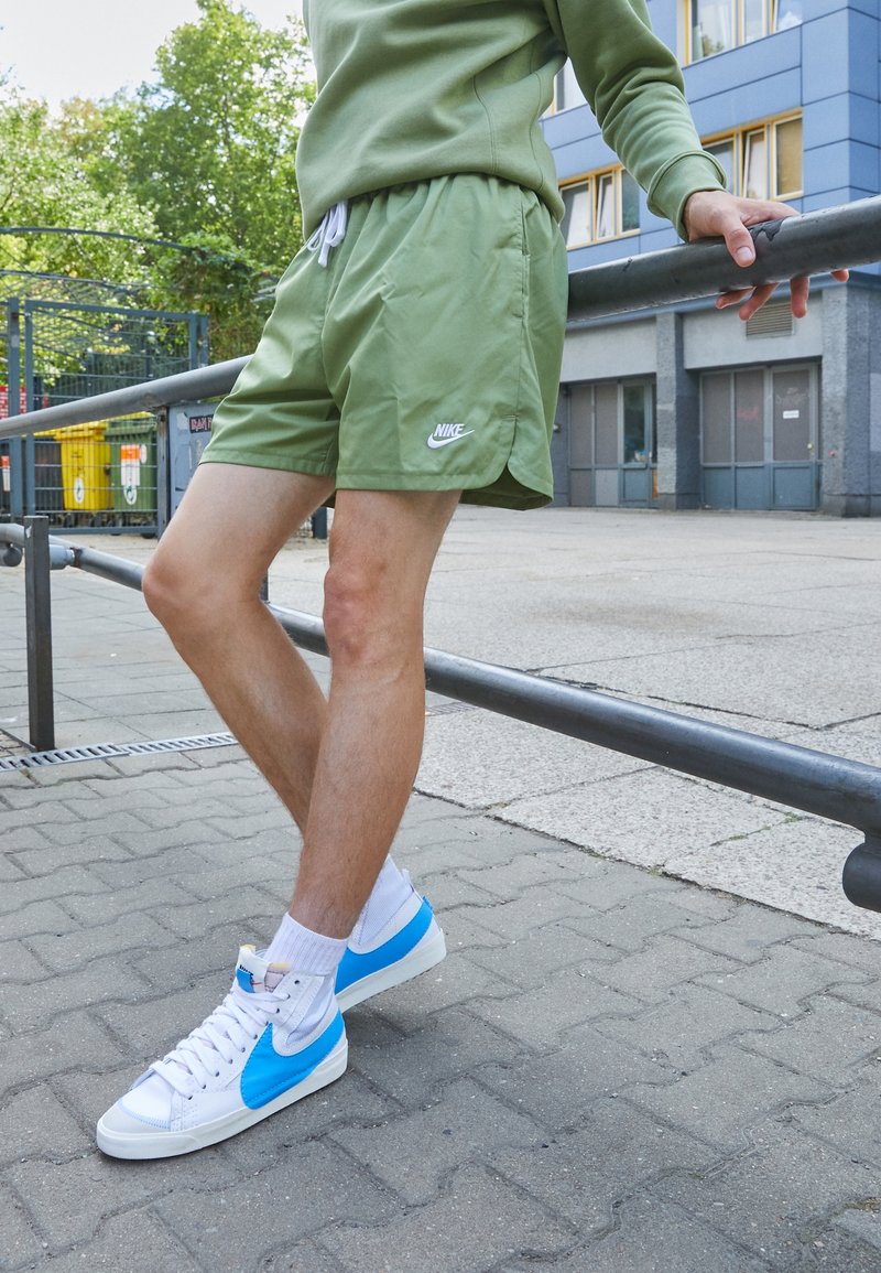 Světlé zelené sportovní šortky s šňůrkou a postranními rozparky, doplněné bílou a modrou obuví Nike ve stylu vysokých tenisek. Texturované asfaltové pozadí.
