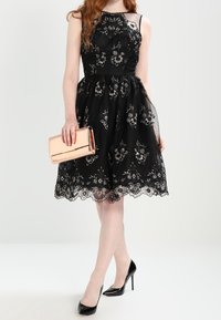 Černé krajkové šaty s květinovou výšivkou, přiléhavým živůtkem a rozšiřující se sukní. Doplněné o kovové psaníčko a černé vysoké podpatky.