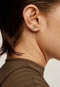 Pendientes de plata con tres pequeños acentos redondos, colocados muy juntos, llevados en el lóbulo de la oreja sobre un fondo marrón claro.