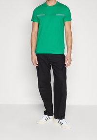 Zielona bawełniana koszulka z okrągłym dekoltem i krótkimi rękawami, z poziomymi szarymi paskami na klatce piersiowej, w połączeniu z czarnymi dżinsami.