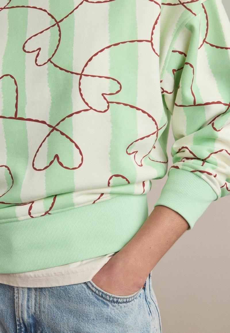 Pull avec des rayures verticales vert clair et crème, motif de lignes en forme de cœur rouge, porté avec un jean bleu clair, main dans la poche.