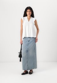 Top in cotone bianco con maniche corte e dettagli ad occhio, abbinato a una lunga gonna di denim. Scarpe nere piatte e una piccola borsa completano l'outfit.