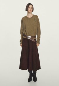 Donna con capelli corti e scuri, che indossa un maglione a V verde oliva, una spilla argentata, una cintura larga marrone, una lunga gonna scura e stivali neri, in piedi.
