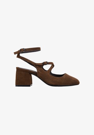 Zapatos de ante marrón con tacón cuadrado y punta afilada, que presentan una correa en el tobillo y un diseño recortado, acabado texturizado y detalle de hebilla ajustable.