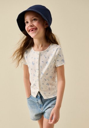Smilende pige iført en marineblå bøllehat, hvid kortærmet blomstret skjorte med knapper og cowboyshorts med hænderne i lommerne.