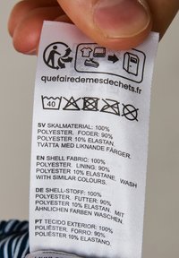 Valkoinen kangasmerkki mustalla tekstillä. Luettelee materiaalin sisällöt: 100 % polyesteri, 90 % polyesterivuori ja 10 % elastaania. Sisältää hoitosymbolit.