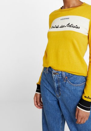 Osoba v žlutém svetru s bílým pruhem a černým nápisem "Entrée des Artistes", v kombinaci s modrými džíny s vysokým pasem a černými zápěstními pásky.