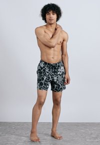 Muž bez trička stojí naboso na šedom podlahe, oblečený v plavkách s čiernymi a bielymi kvetinovými vzormi, dotýkajúci sa svojho ľavého ramena.