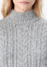 Nahaufnahme einer Person, die einen hellgrauen, grob gestrickten Pullover mit Kabelmustern und einem gerippten hohen Kragen trägt.