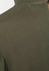 Tissu léger vert olive avec une texture lisse. Doté d'un col montant et d'un détail zip à l'arrière. Design décontracté.