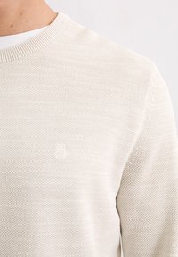 Jasnobeżowy, dziergany sweter z okrągłym dekoltem, o subtelnej fakturze i małym, wytłoczonym logo na piersi. Gładkie wykończenie tkaniny.