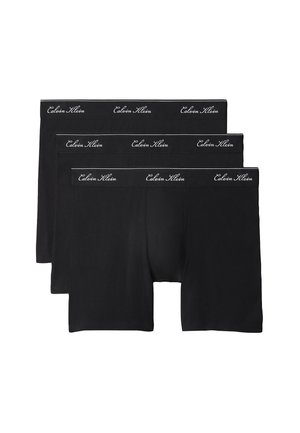 Hemdloser Mann mit lockigem Haar, der schwarze Boxershorts trägt, barfuß vor einem schlichten grauen Hintergrund steht.
